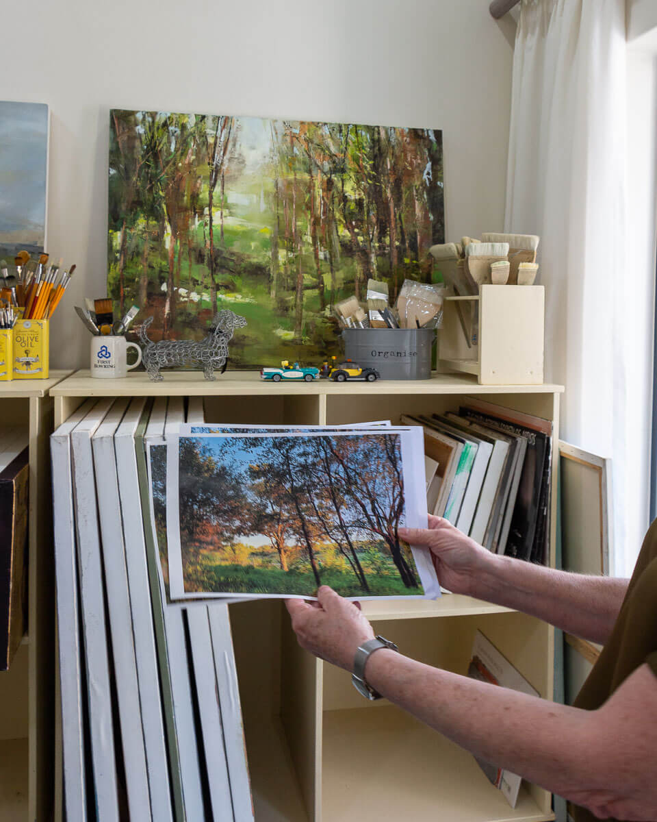 Janet Dirksen's hand holding photographs of a forest landscape in the foreground, with her painting depicting a forest environment in the background. The photos and painting are in shades of brigh green and brown.